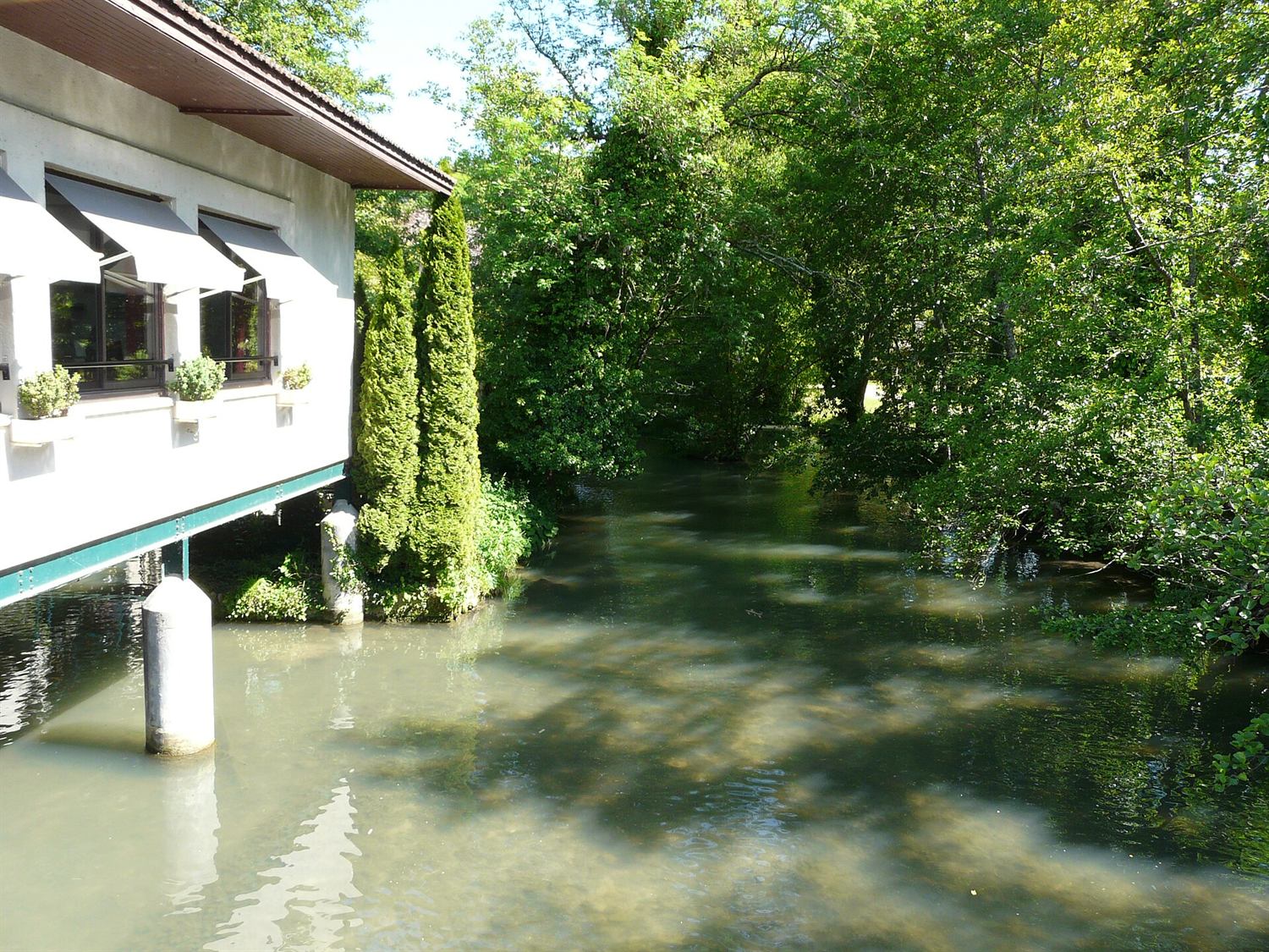 La rivière Couze, aux eaux vertes et calmes, longeant un bâtiment sur pilotis au milieu des arbres.