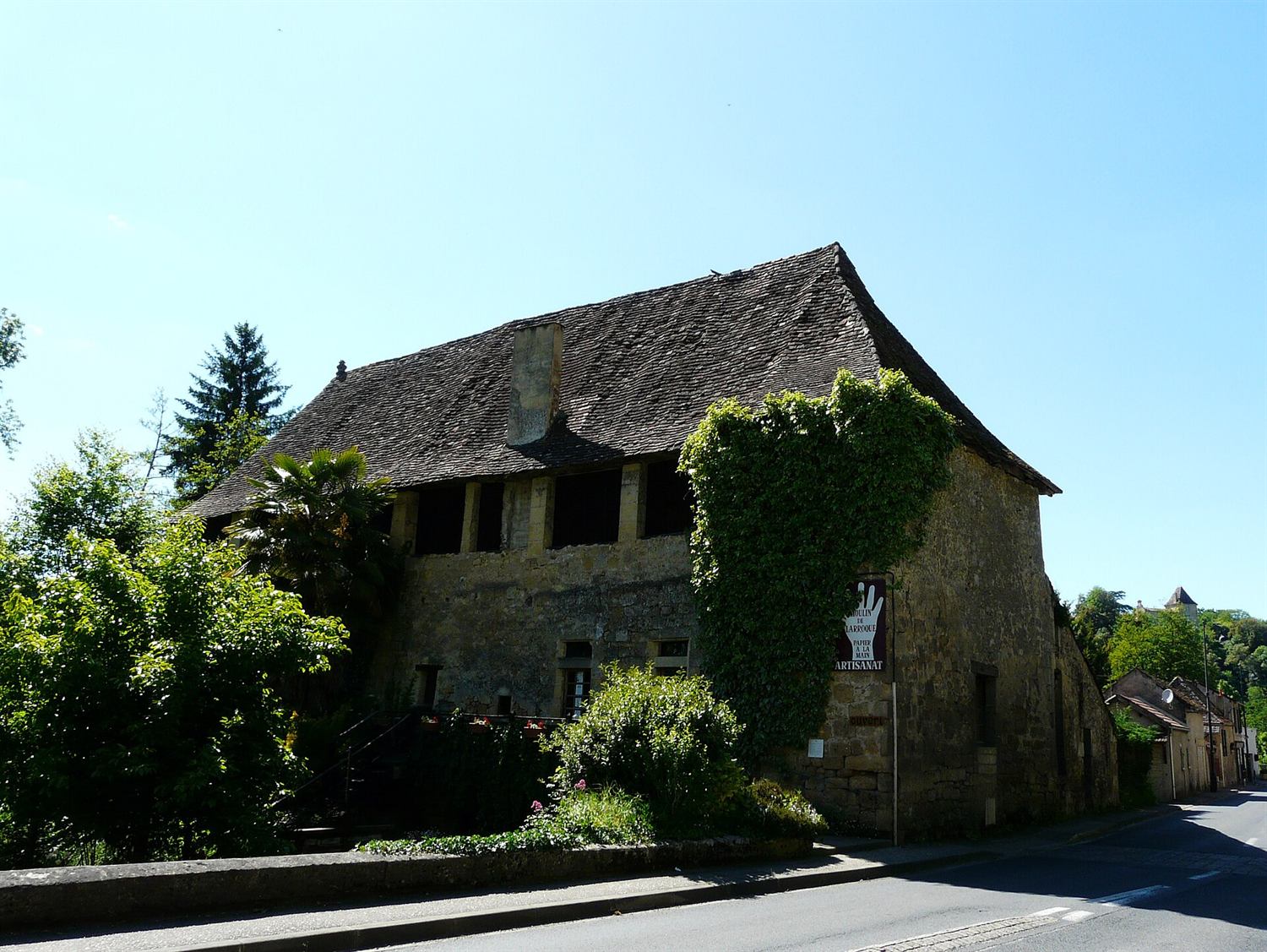 Le moulin de Larroque, grand bâtiment de pierre au toit de tuiles brunes couvert de lierre, en bord de route.